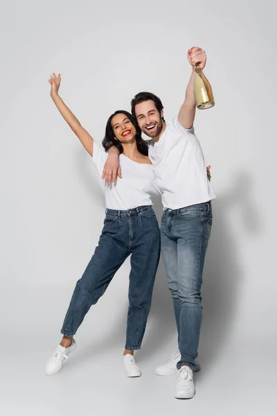 Full length view of young man holding champagne bottle in raised hand near happy woman on grey — Stock Photo