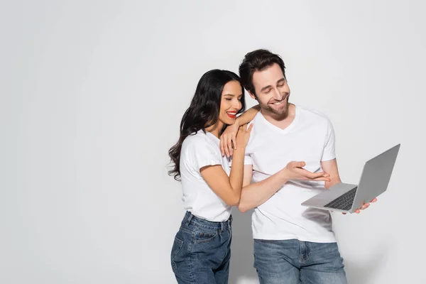 Homme souriant en t-shirt blanc et jeans pointant vers ordinateur portable près petite amie heureuse sur gris — Photo de stock