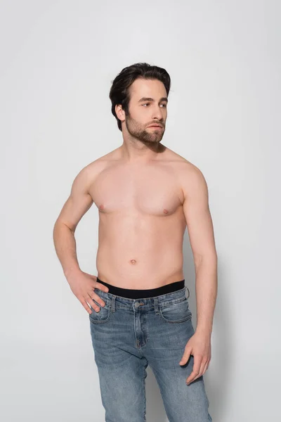 Без сорочки чоловік в джинсах дивиться геть, стоячи з рукою на стегні на сірому — стокове фото