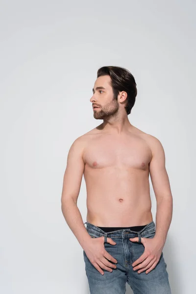 Brunette shirtless man in jeans looking away while standing with thumbs in belt loops on grey — Stock Photo