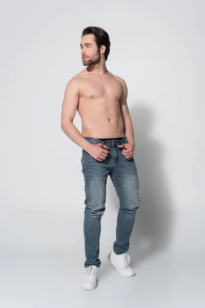 Full length view of young shirtless man in jeans holding thumbs in belt loops while looking away on grey — Stock Photo