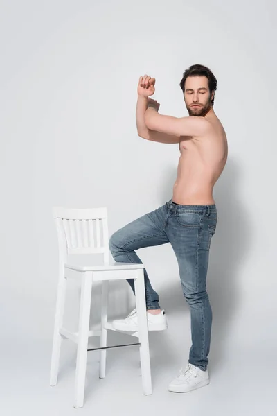 Full length view of shirtless man in jeans posing near white chair on grey — Stock Photo