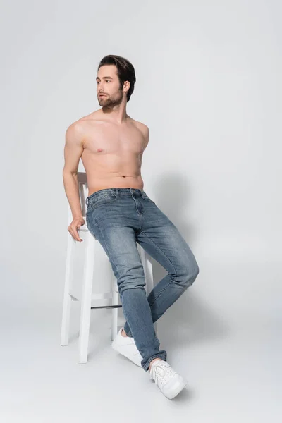 Vista completa de hombre sin camisa en jeans mirando hacia otro lado mientras posando en silla blanca en gris - foto de stock