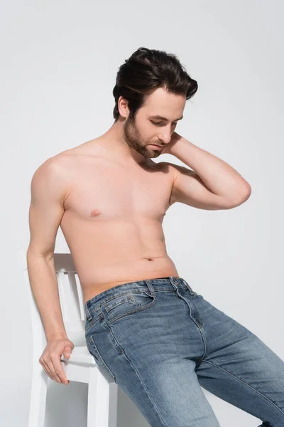 Uomo senza maglietta in jeans toccare il collo mentre seduto sulla sedia bianca su grigio — Foto stock