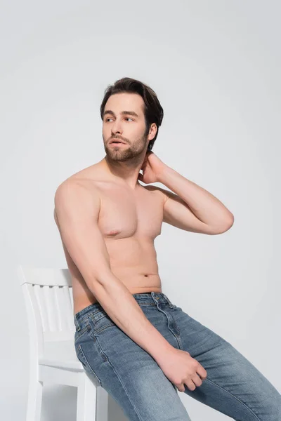 Sexy shirtless man in jeans touching neck and looking away on white chair on grey — стоковое фото