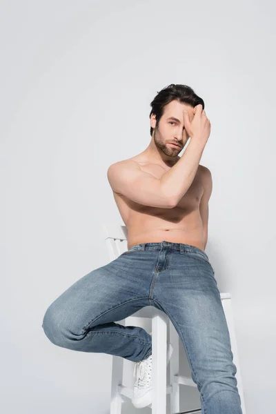 Brunette shirtless man in jeans looking at camera while holding hand near face on grey — Stock Photo