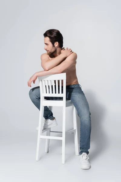 Vue pleine longueur de l'homme torse nu sexy en jeans assis sur une chaise blanche et regardant loin sur gris — Photo de stock