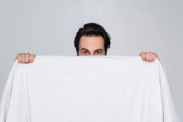 Jeune homme regardant la caméra tout en obscurcissant le visage avec couverture blanche isolée sur gris — Photo de stock