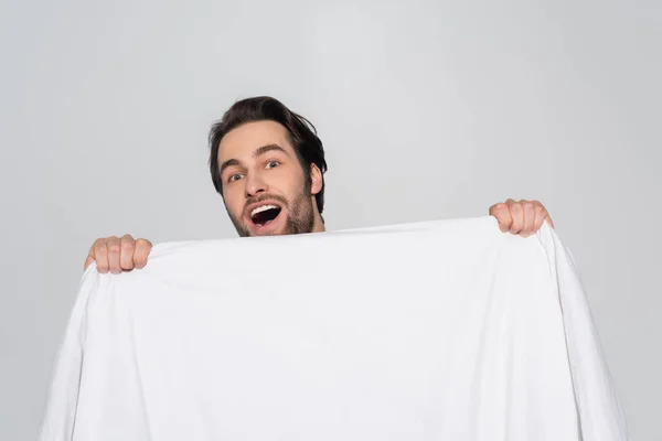 Amazed brunette man looking at camera behind white blanket isolated on grey — Stock Photo