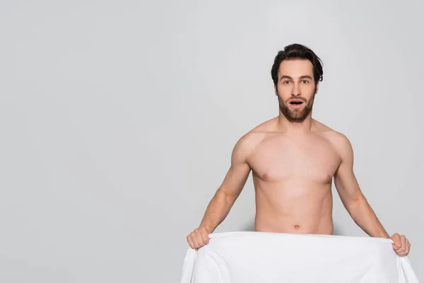 Homem sem camisa surpreso segurando cobertor branco enquanto olha para a câmera no cinza — Stock Photo