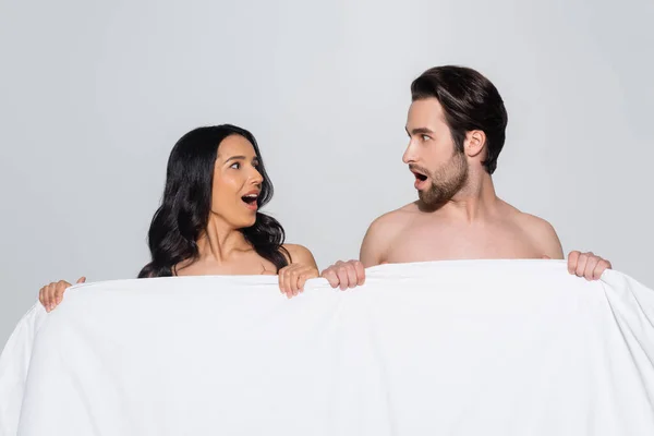 Erstaunt schauen Mann und Frau einander an, während sie eine weiße Decke isoliert auf grau halten — Stockfoto