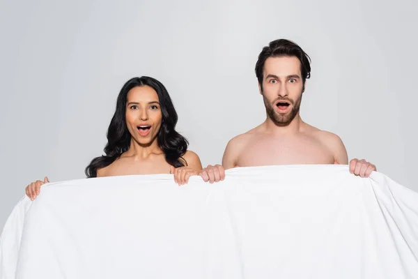 Asombrado hombre y mujer sin camisa con hombros desnudos mirando a la cámara cerca de manta blanca aislada en gris - foto de stock