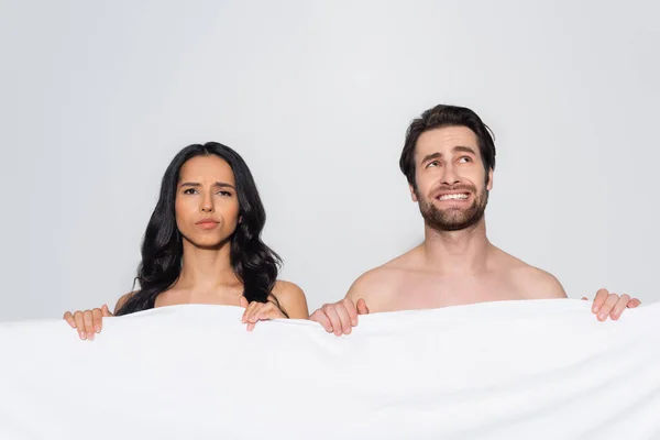 Недовольная женщина смотрит в камеру рядом с смущенным мужчиной и белым одеялом, изолированным на сером — стоковое фото