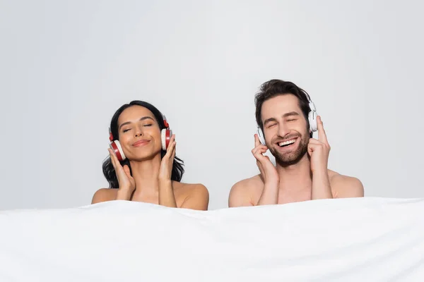 Freudiges Paar mit geschlossenen Augen, das Musik in Kopfhörern in der Nähe einer weißen Decke auf grau isoliert hört — Stockfoto