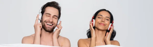 Lächelndes Paar mit geschlossenen Augen, das Musik in Kopfhörern in der Nähe einer weißen Decke genießt, isoliert auf grau, Banner — Stockfoto