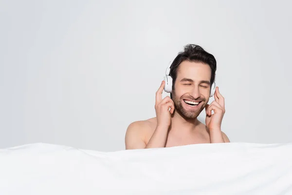 Мужчина без рубашки с закрытыми глазами улыбается, слушая музыку в наушниках за белым одеялом, изолированным на сером — стоковое фото
