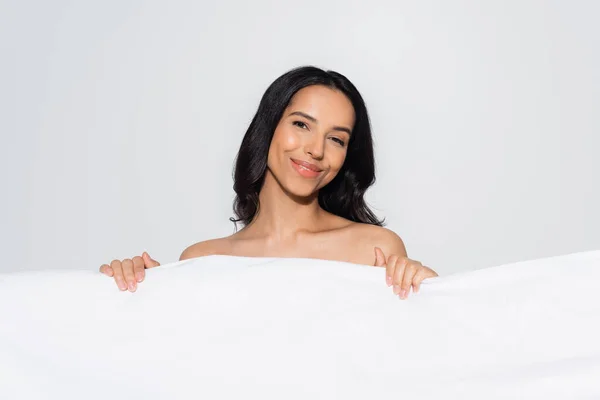 Morena mujer sonriendo a la cámara mientras posando detrás de una manta blanca aislada en gris - foto de stock