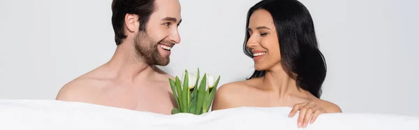 Homme torse nu heureux regardant femme souriante près de couverture blanche et tulipes isolées sur gris, bannière — Photo de stock