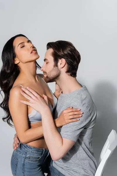 Чуттєва брюнетка жінка з закритими очима біля чоловіка в футболці, обіймаючи її на сірому — стокове фото