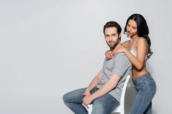 Heiße Frau in BH und Jeans umarmt jungen Mann auf Stuhl sitzend und schaut in die Kamera auf grau — Stockfoto