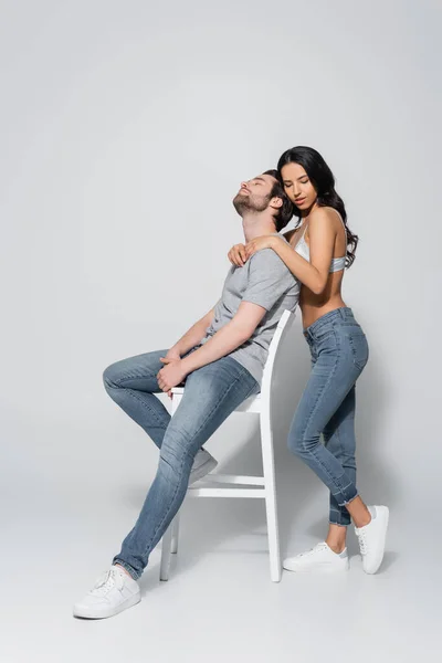 Full length view of brunette woman in jeans and bra hugging man sitting on chair on grey — Stock Photo