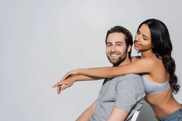 Sensual brunette woman in bra hugging man smiling at camera while sitting on grey — Stock Photo