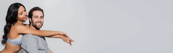 Young man smiling at camera near seductive woman in bra embracing him isolated on grey, banner — Stock Photo