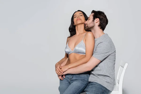 Homme en t-shirt étreignant femme passionnée en soutien-gorge satin assis sur chaise sur gris — Photo de stock