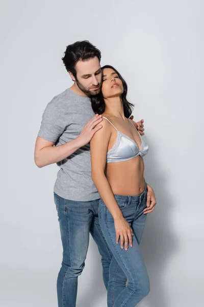 Homme en t-shirt et jeans étreignant femme sexy en soutien-gorge debout avec les yeux fermés sur gris — Photo de stock