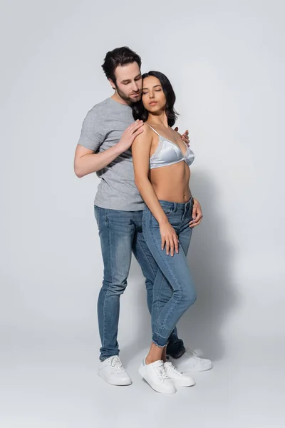 Full length view of man embracing woman in jeans and bra on grey — Stock Photo