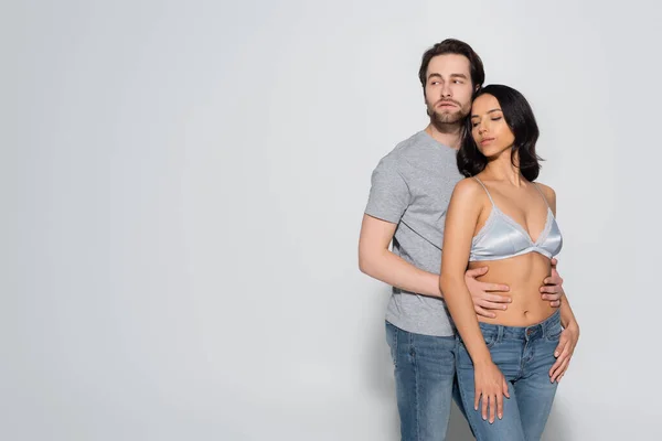 Young man in t-shirt looking away while hugging hot woman in satin bra on grey — Stock Photo