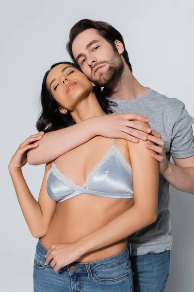 Young man looking at camera while hugging woman in satin bra on grey — Stock Photo