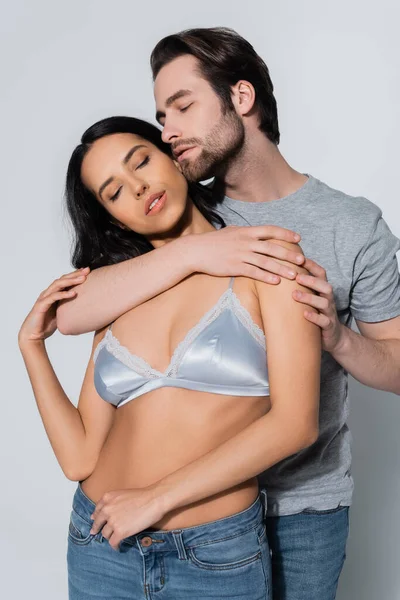 Mujer caliente en sujetador de satén de pie con los ojos cerrados cerca de hombre joven seduciéndola en gris - foto de stock