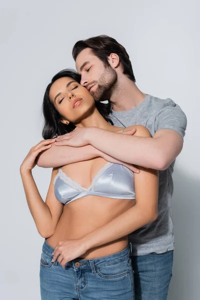 Молодой человек обнимает сексуальную девушку в атласном лифчике стоя с закрытыми глазами на сером — стоковое фото