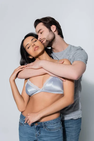 Jeune homme étreignant fille sexy en soutien-gorge satin debout avec les yeux fermés sur gris — Photo de stock