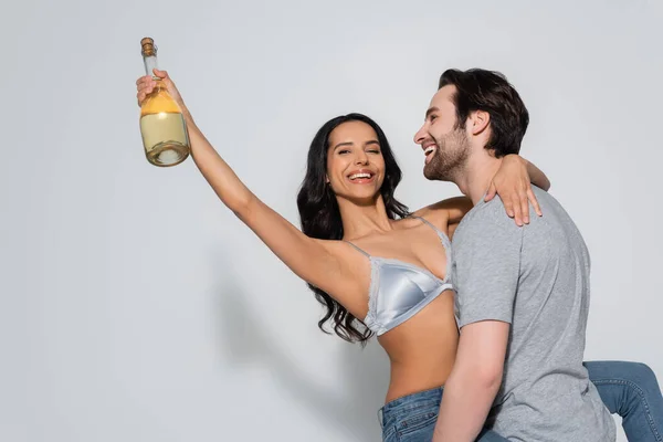 Щасливий чоловік у футболці тримає сексуальну жінку з пляшкою шампанського на сірому — стокове фото