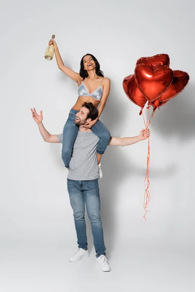 Happy man with red balloons piggybacking woman in bra holding champagne bottle on grey — Stock Photo