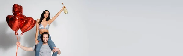 Uomo con palloncini rossi a cavalletto donna felice in reggiseno che tiene champagne sul grigio, banner — Foto stock