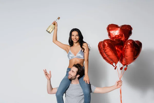 Улыбающийся мужчина с воздушными шарами в форме сердца, поддерживающий женщину, держащую шампанское на сером — стоковое фото
