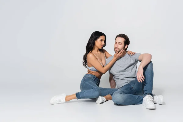 Full length view of passionate woman in jeans and bra touching face of young man while sitting on grey — Stock Photo