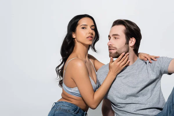 Femme brune passionnée en jeans et soutien-gorge touchant le visage du jeune homme tout en regardant la caméra isolée sur gris — Photo de stock