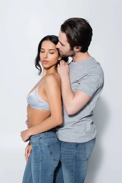 Hombre en camiseta abrazando a mujer seductora en sujetador y tocándose el pelo en gris - foto de stock