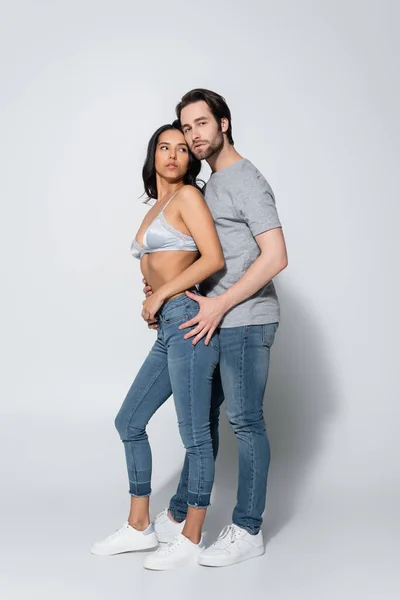 Ansicht des brünetten Mannes in voller Länge, der in die Kamera schaut, während er in der Nähe einer sexy Frau in Jeans und BH auf grau steht — Stockfoto