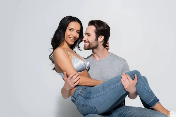 Happy man in t-shirt holding sexy woman in bra looking at camera on grey — Stock Photo