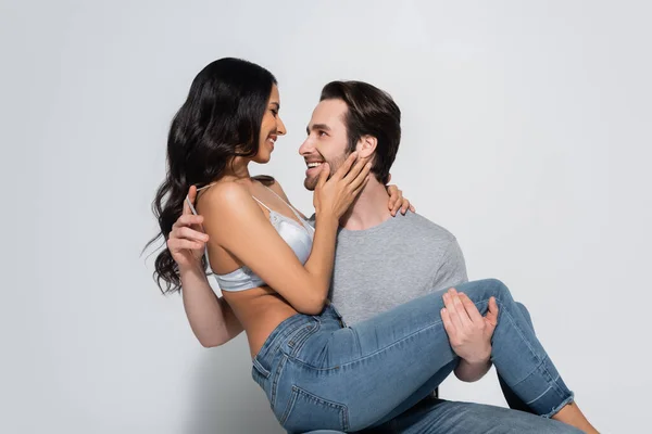 Happy man in t-shirt holding woman in jeans and bra touching his face on grey — Stock Photo