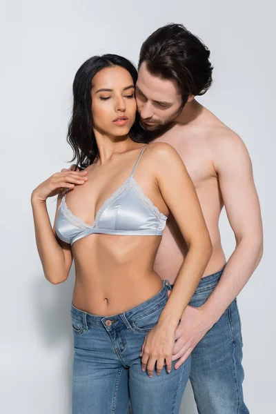 Sexy shirtless man in jeans embracing passionate brunette woman in bra on grey — Stock Photo