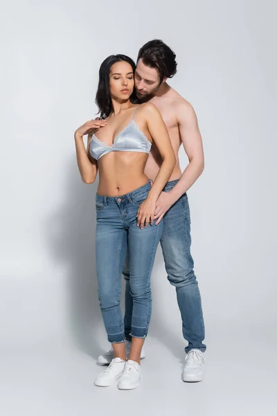 Full length view of shirtless man and sexy woman in bra standing in jeans on grey — Stock Photo