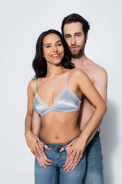 Sexy woman in bra looking at camera while shirtless man holding thumbs in belt loops of her jeans on grey — Stock Photo