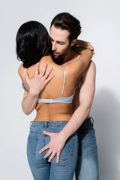 Back view of passionate woman in jeans and bra near young shirtless man hugging her on grey — Stock Photo