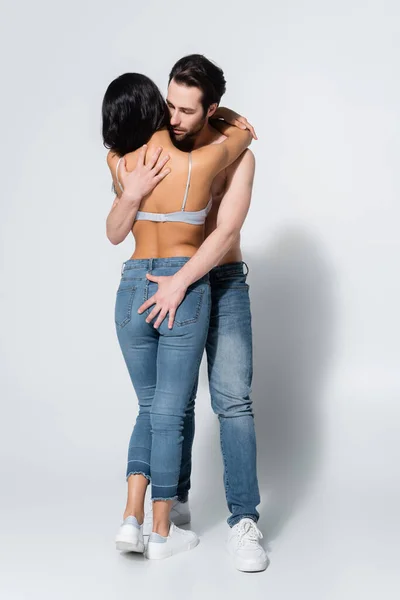 Back view of sexy woman in jeans and bra near shirtless man hugging her on grey — Stock Photo